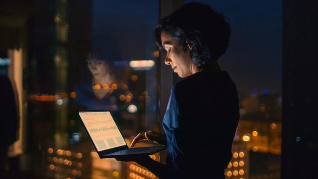 Successful, Stylishly Dressed Businesswoman Holds Laptop While Standing Near the Window of Her Office. Late at Night Professional Woman Doing Important Job. Window Has Big City View with Many Lights