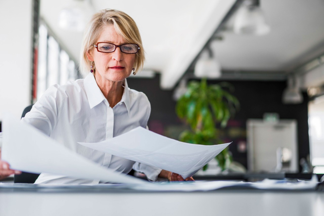 woman with blonde hair and glasses looks through files at a table.