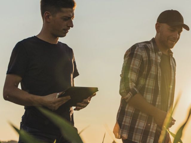 Due persone camminano in un campo coltivato al tramonto: una consulta un tablet e l’altra indossa una camicia a quadri e cappellino