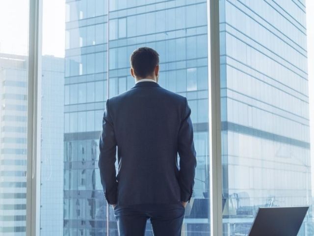 Un manager di spalle osserva i palazzi moderni dalla vetrata del suo ufficio