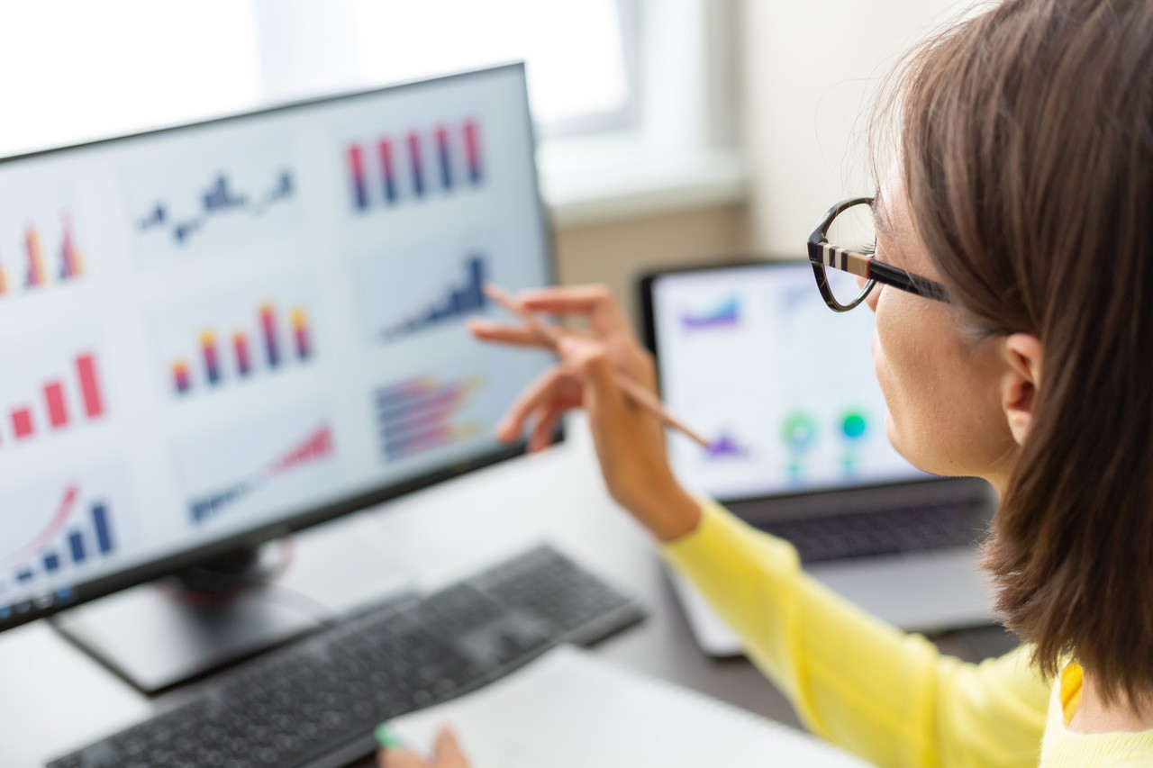 woman in yellow long sleeve and glasses faces a monitor and computer screen filled with different types of graphs with a notebook and pencil in hand.